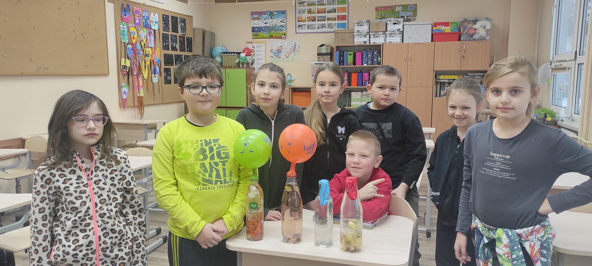 Uczniowie stojący przy stoliku, podczas doświadczenia z butelkami napełnionymi bioodpadami, na których znajdują się napompowane gazem balony.