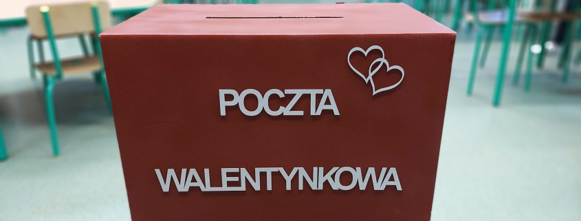 Na zdjęciu znajduje się czerwone pudełko, z napisem " Poczta walentynkowa". Pudełko znajduje się na stoliku z zielonym obrusem, na tle klasy szkolnej.