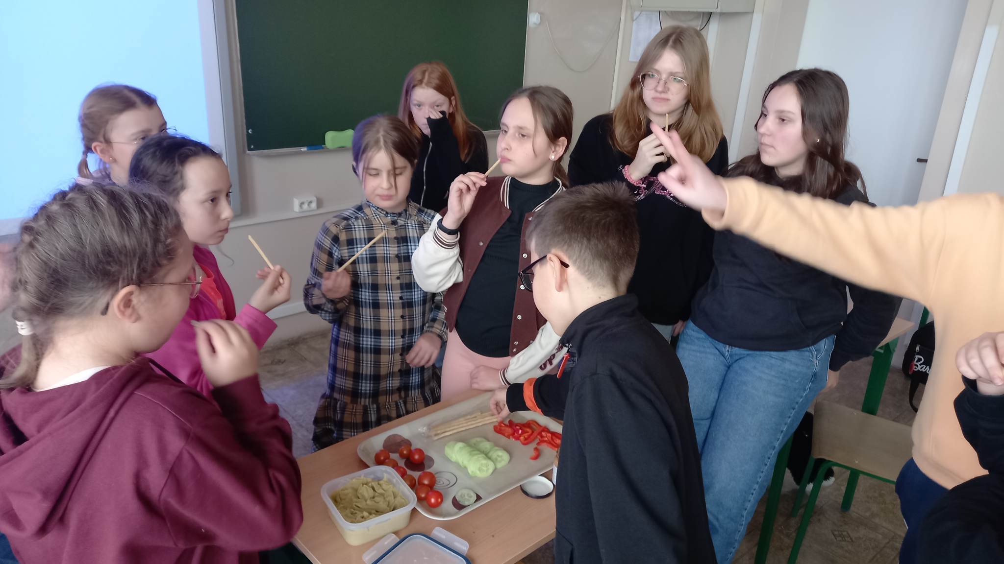 Na zdjęciu widać uczniów stojących w sali lekcyjnej wokół stolika, na którym znajdują się warzywa, hummus i paluszki grissini. Uczniowie degustują przysmaki.