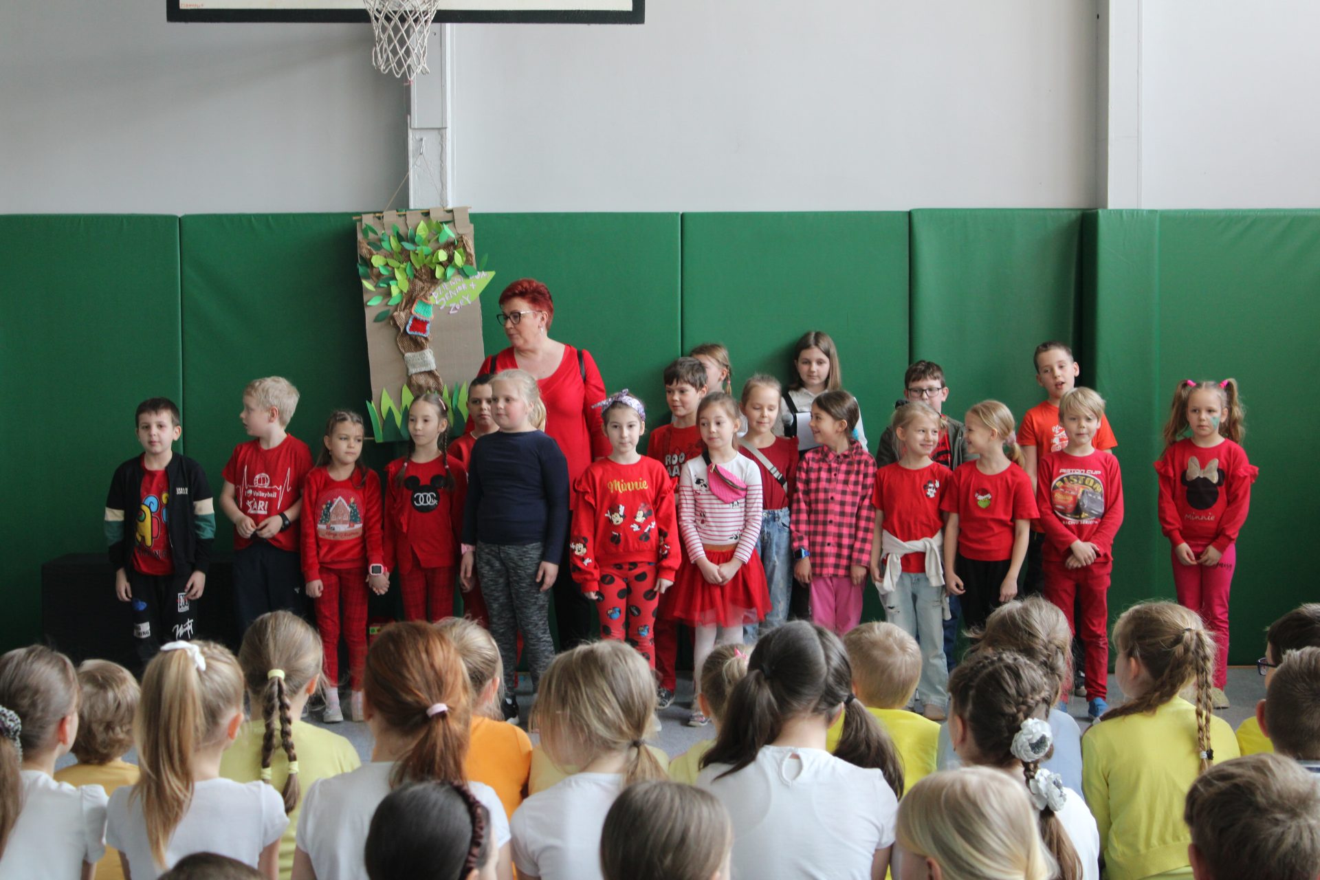 Uczniowie z nauczycielem ubrani na czerwono stoją przed swoimi starszymi kolegami na sali gimnastycznej.