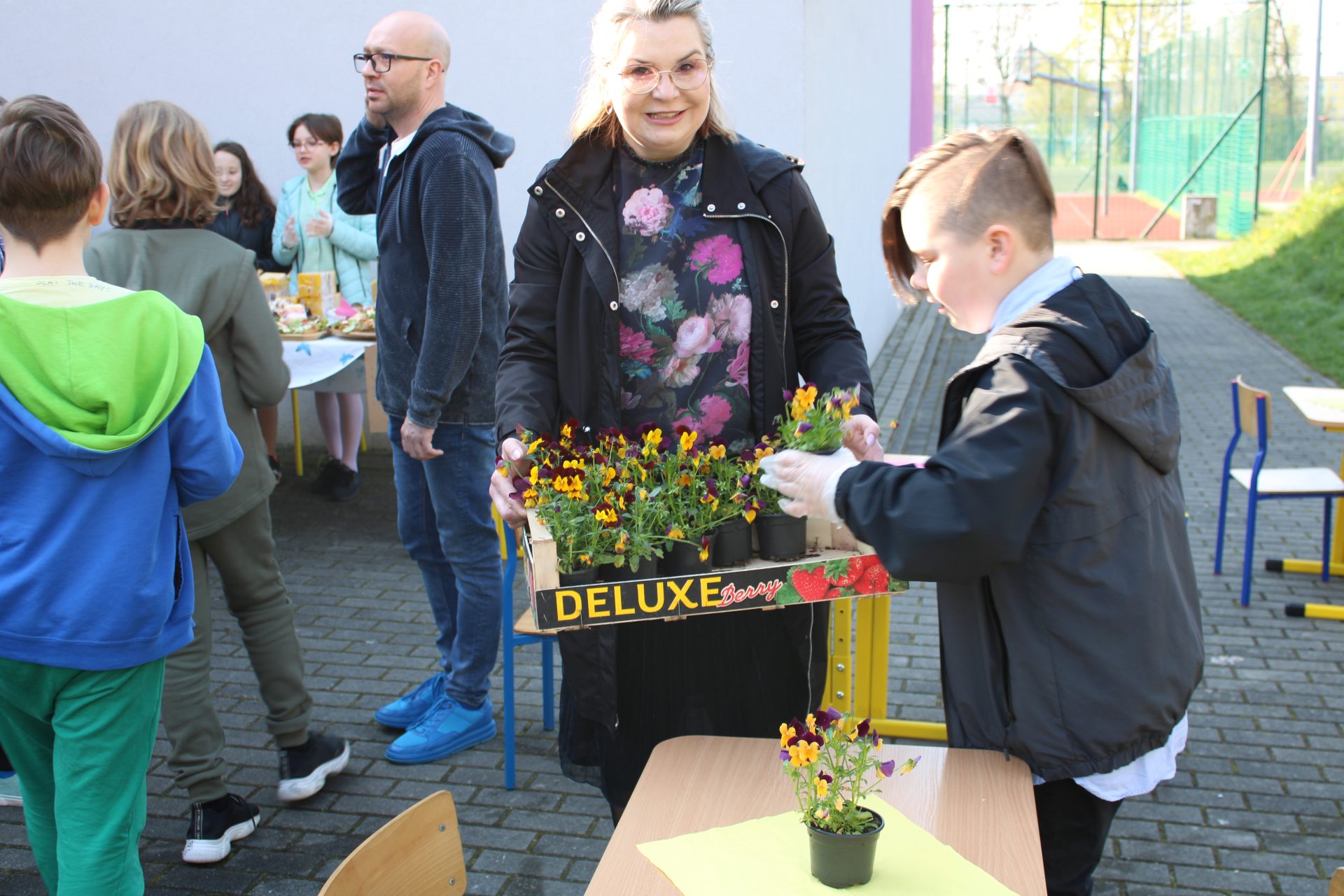 nauczycielka trzyma skrzynki z bratkami. Obok uczeń wyjmuje doniczki z kwiatkami. W tle uczniowie oraz kawiarenka szkolna.