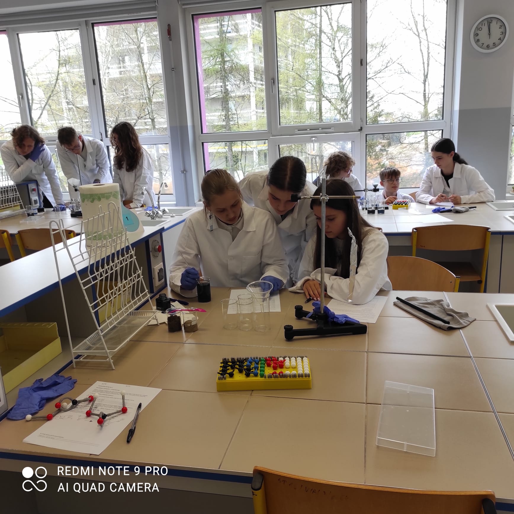 Uczestnicy w białych fartuchach wykonują doświadczenia z zakresu fizyki, biologiii chemii.