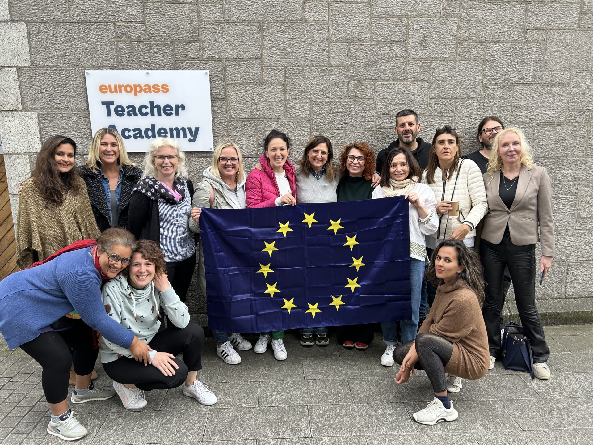 Na tle ściany budynku z napisem europass Teacher Academy stoi 14 uczestników kursu wraz z prowadzącym. Osoby w środku trzymają flagę Unii Europejskiej.