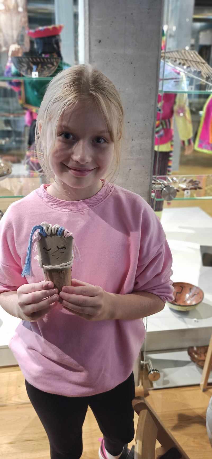  Lalka z upcyklingu – na zdjęciu widać uśmiechniętą dziewczynkę w różowej bluzie, która trzyma w ręku własnoręcznie wykonaną laleczkę. W tle muzealne gabloty z ekspomatami.
