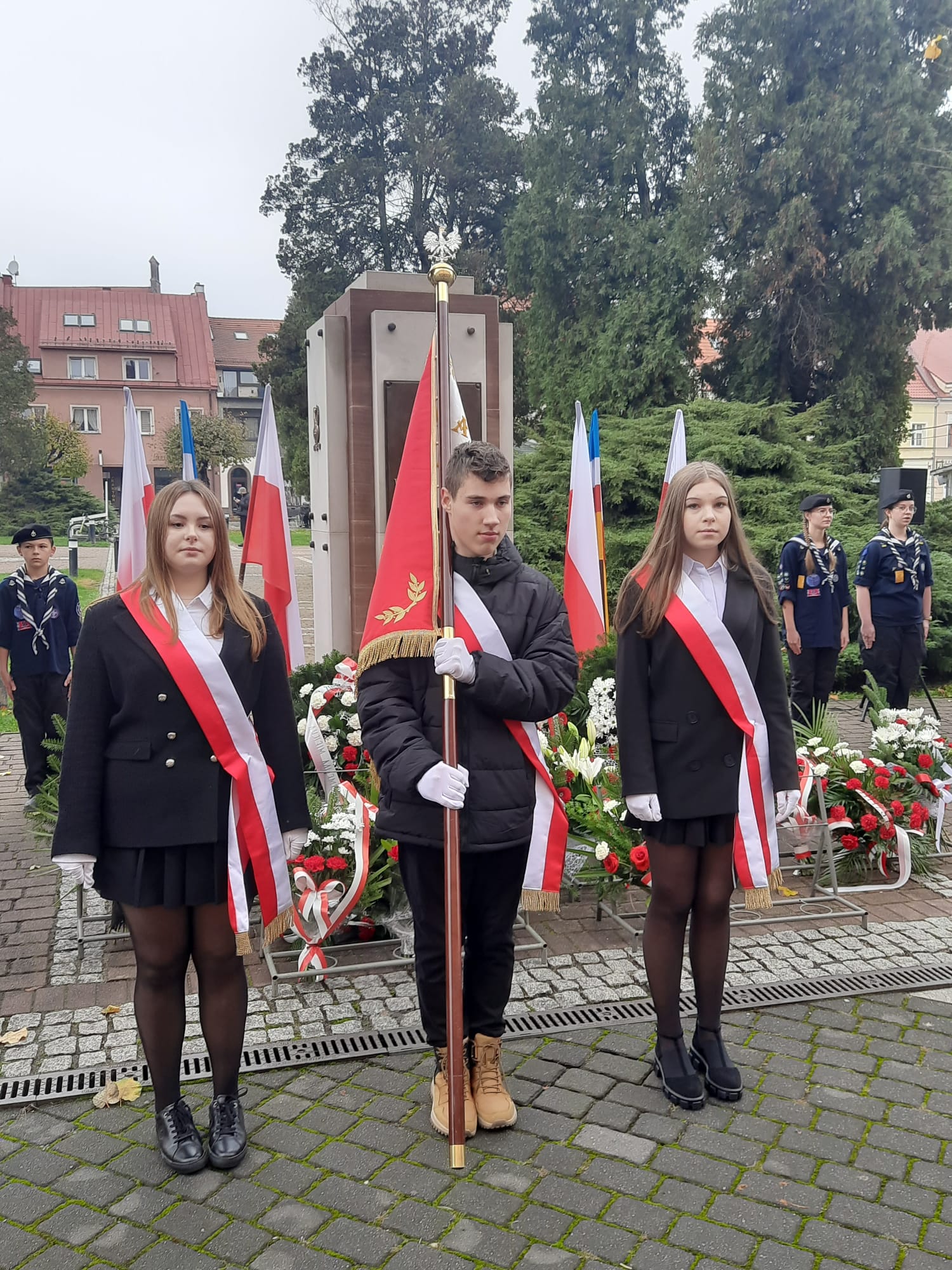 Na zdjęciu widać poczet sztandarowy na tle pomnika Pamięci Walczących o Wolną Polskę. Chorąży trzyma sztandar szkoły.