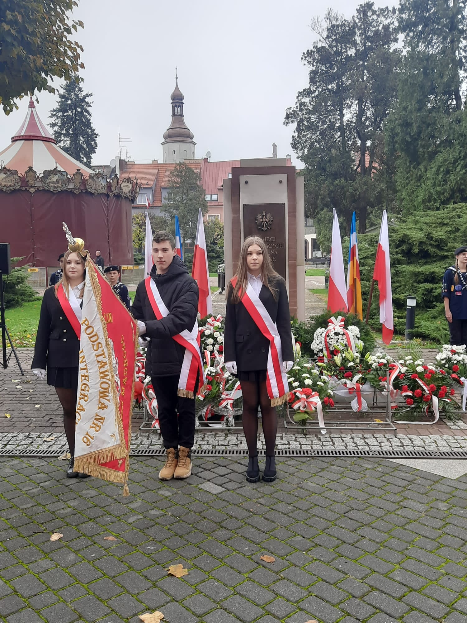 Na zdjęciu widać poczet sztandarowy na tle pomnika Pamięci Walczących o Wolną Polskę. Chorąży trzyma pochylony sztandar szkoły.