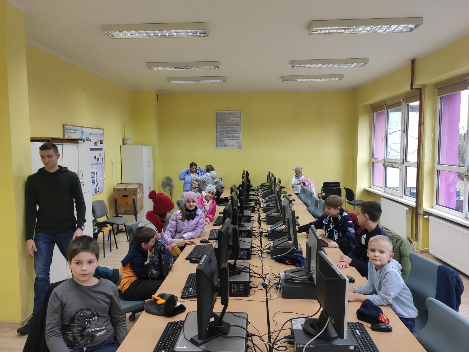 Pracownia komputerowa, przy stanowiskach siedzą chłopcy i dziewczynki z lewej strony opiekun.