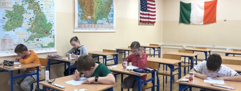 Zdjęcie przedstawia uczniów klas 3 siedzących w ławkach i rozwiązujących zadania matematyczne. W tle na ścianach mapy oraz flagi.